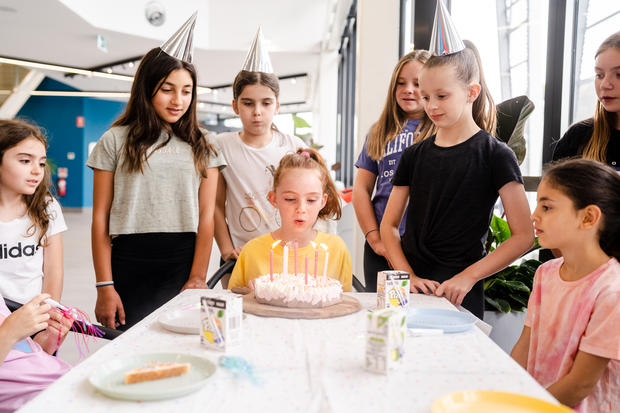 Group of eight young girls celebrating a birthday. Each are surround a young girl in a yellow tee shirt who is blowing out birthday candles