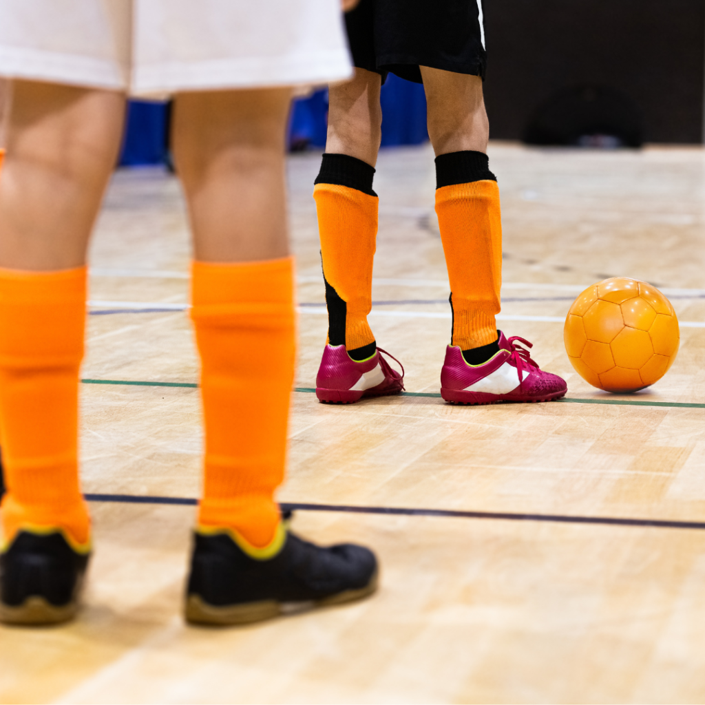 Photograph of two players on stadium court. Foreground player is wearing orange socks and black shoes. Background player is wearing red shoes and is placed to kick the orange ball.